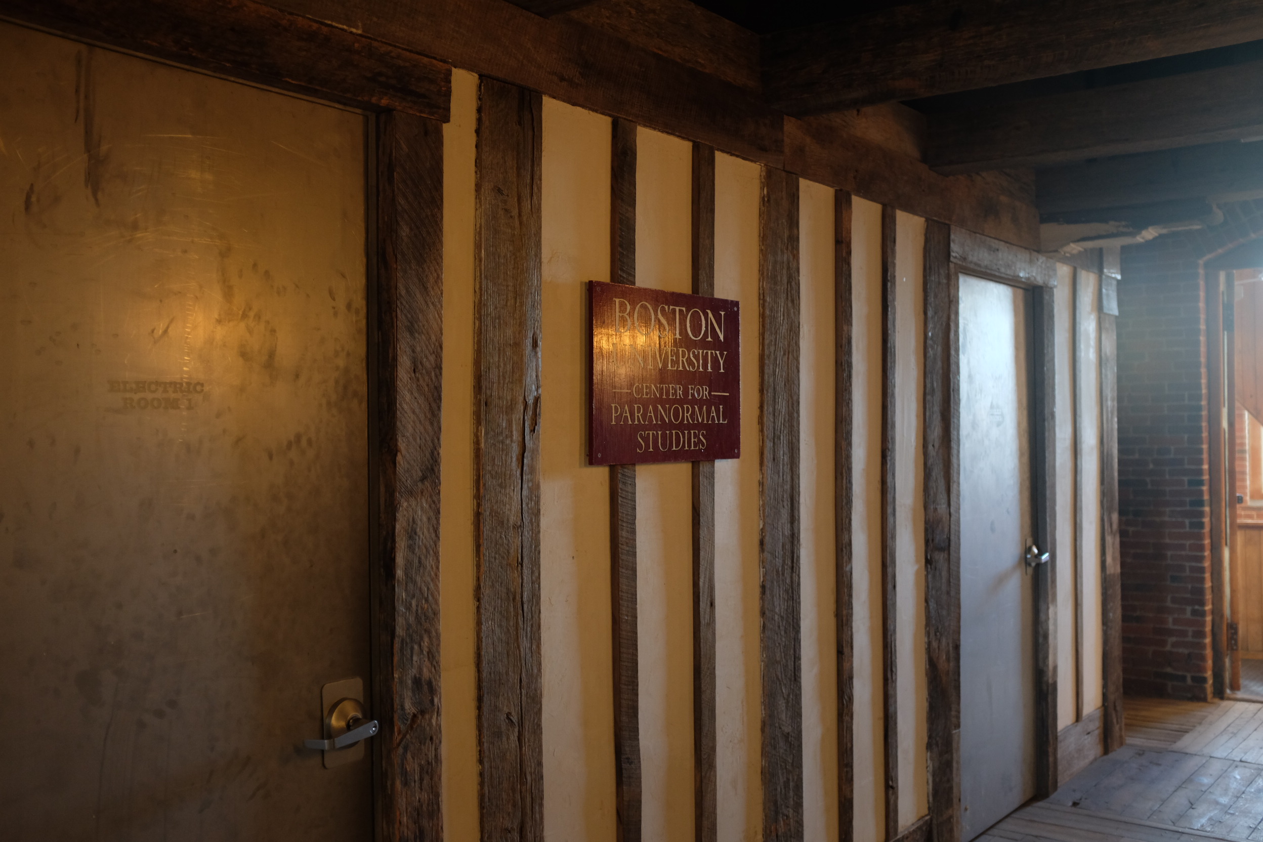 A photo from an angle of an old wooden wall which is painted yellow; a sign in the center reads 'Boston University Center for Paranormal Studies'.
