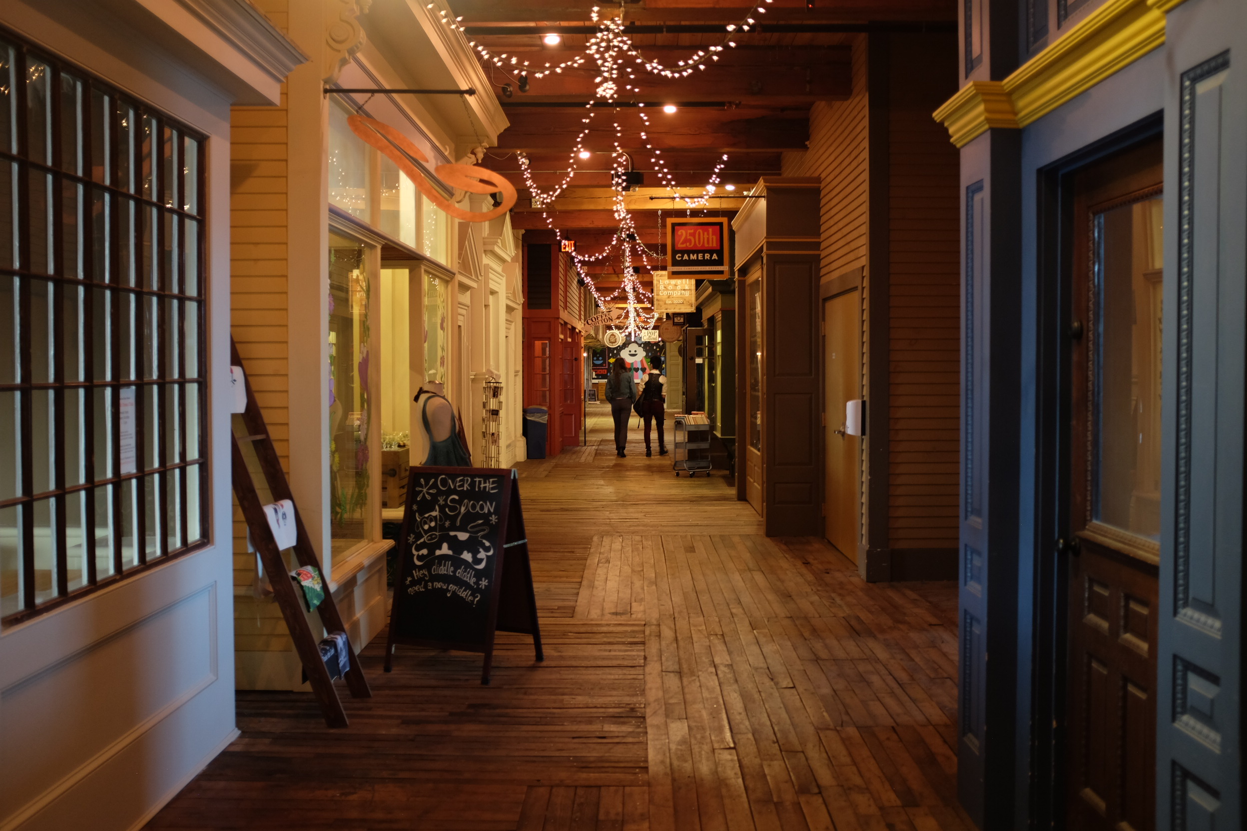 A photo taken down a hallway lined with old-fashioned storefronts and lit by incandescent string lights hanging from the ceiling; Two people are walking away from the camera.