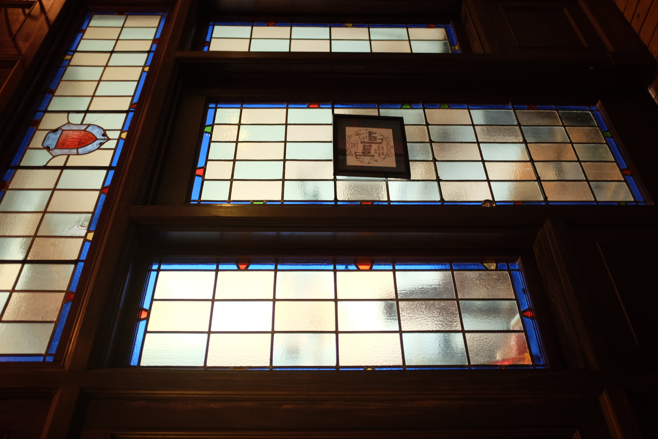 A photo from an angle of four stained glass panels, three in a row with a fourth across the top. All three panels are checkered in blue and pink. The top panel has a shield emblem in red, blue, and green. The bottom middle panel has a sign which reads 'Tone Loft'.