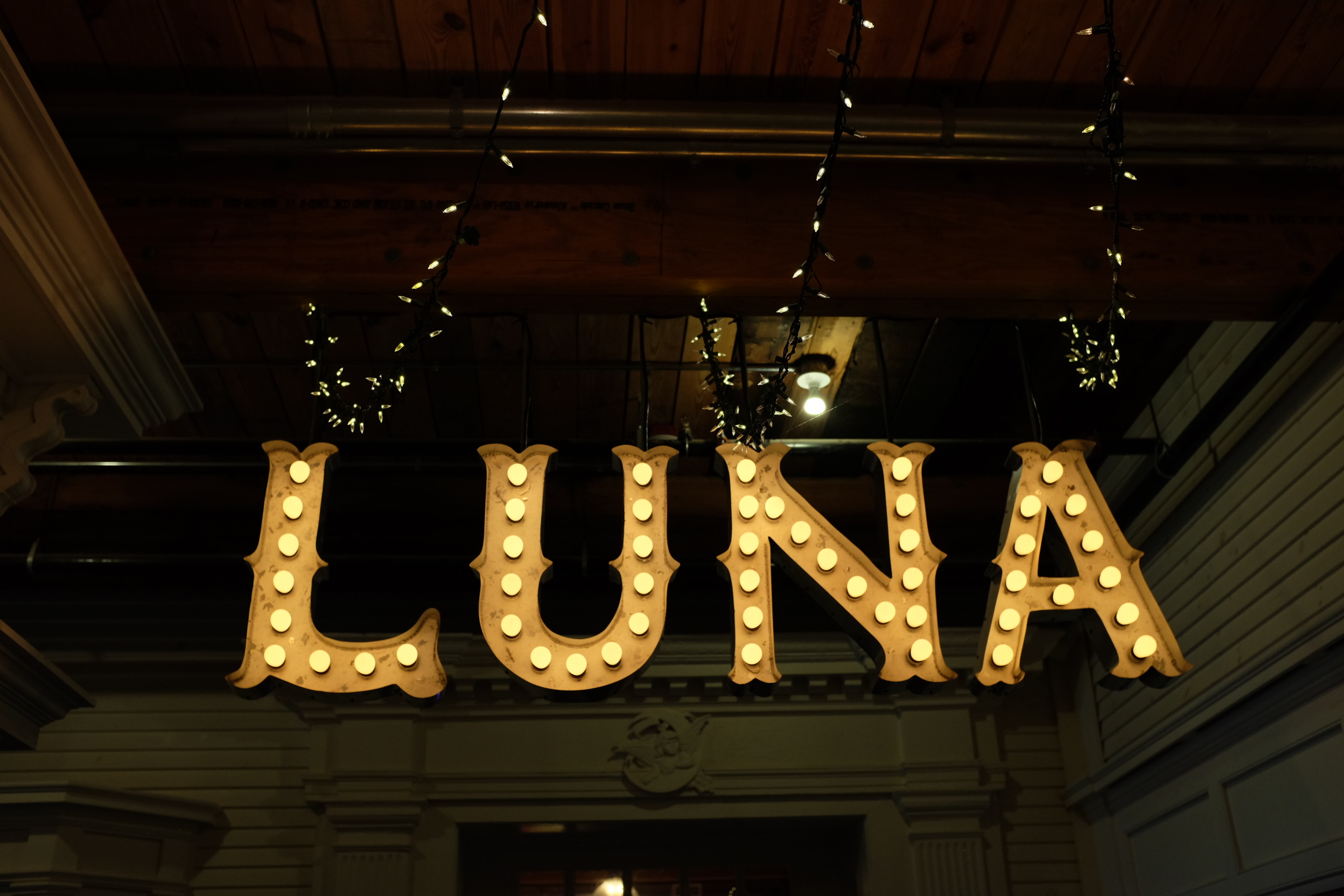 A photo of a sign suspended from the ceiling which reads 'Luna' with incandescent lightbulbs tracing the letters.